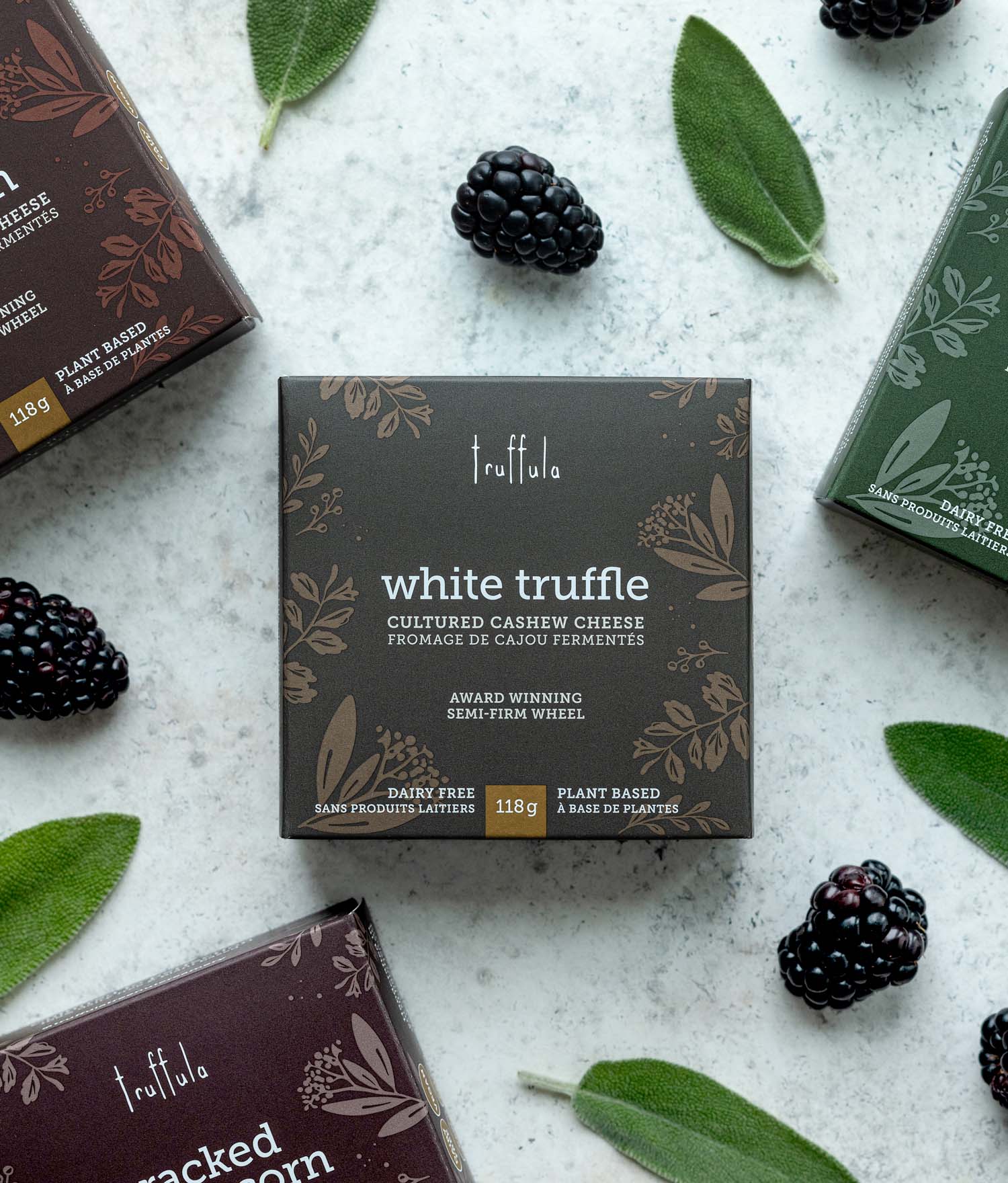 Brown square cheese box with text 'White Truffle' on the front, on white background, surrounded by fresh sage leaves and blackberries.
